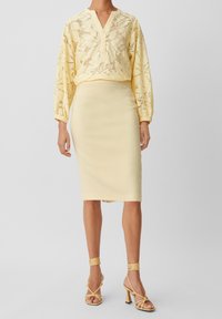 Kvinna i en gul spetsblus med blommönster, en figursydd knälång pennkjol och sandaler med hög klack och remmar.