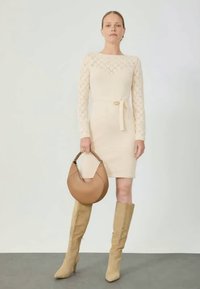 Vestido de malha em bege claro com mangas longas de renda, cintura ajustada e botas de joelho em bege combinando; segurando uma bolsa meia-lua em tom açucarado.