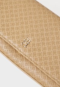 Beige teksturert lærclutch med preget geometrisk mønster og en metallisk logoaccents. Har en strømlinjeformet, rektangulær form.