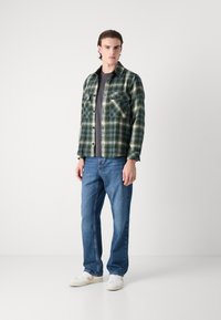 Green checkered flannel shirt with two chest pockets, worn over a gray top, paired with blue straight-leg jeans and white sneakers.