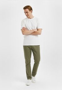 Bijela majica kratkih rukava u kombinaciji s maslinastozelenim hlačama i bijelim tenisicama. Model stoji s prekriženim rukama, smiješi se, nasuprot jednostavne pozadine.