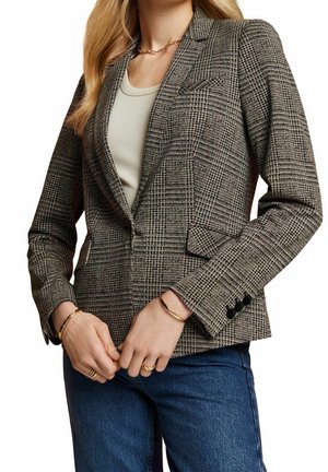 Donna che indossa un blazer a quadri con bottoni neri, canotta bianca, jeans blu, gioielli d'oro, e con le mani congiunte in vita.
