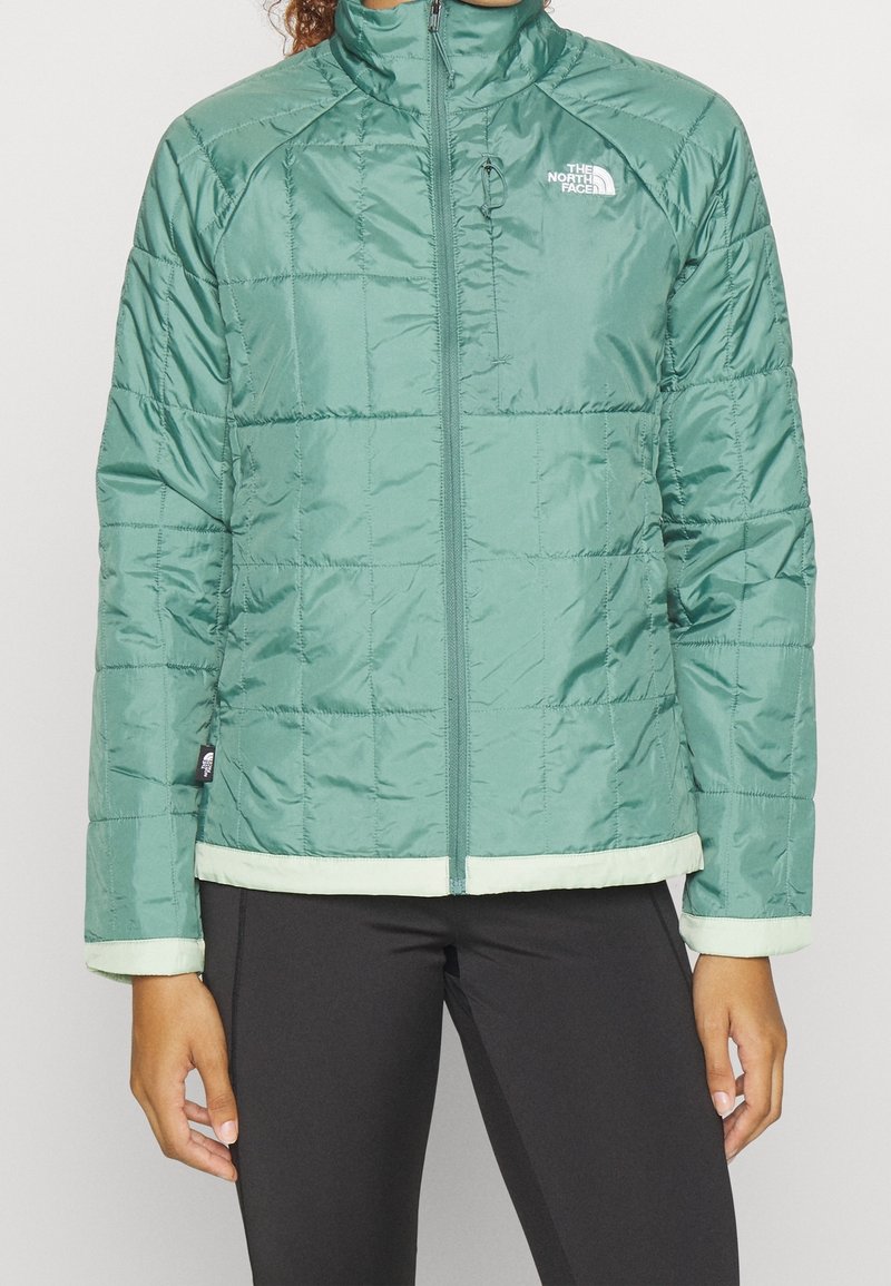 Zelená zateplená bunda s prošívaným vzorem, vysokým límcem, předním zipem a kontrastním světlým zeleným lemem na dolním okraji a manžetách.