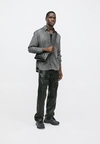 Grå rutete skjorte, svart singlet, svarte cargo-bukser i skinn, svarte joggesko og en svart veske med preget logo, stående mot en lys bakgrunn.