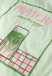 Grün gestreifter Stoff mit einer Grafik eines grünen Matcha-Latte in einem hohen Glas mit einem pink gestreiften Strohhalm und dem Text „Matcha mornings“ darüber.