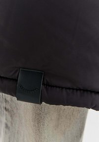 Čierna prešívaná tkanina s prešitým okrajom a malou čiernou etikettou s krivkou a vertikálnymi čiarami pripomínajúcimi riasy.