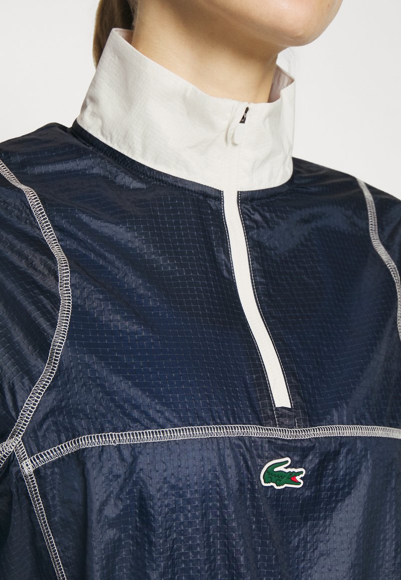 Gros plan sur une personne portant une veste zippée bleu marine et blanche avec des coutures visibles et un petit logo crocodile vert sur la poitrine.