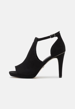 Sort ruskind sandal med høj hæl, åben tå, ankelrem med spænde og en tynd stiletthæl på en hvid baggrund.