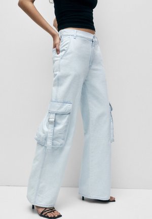 Vrouw draagt lichtblauwe wijde cargobroek met gespdetail en zwarte sandalen met hak en bandjes, staand tegen een effen achtergrond.