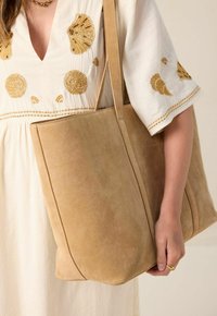 Beige suède tote bag met een gestructureerde vorm en dubbele hengsels, voorzien van een gladde textuur en een minimalistisch ontwerp. Gouden accenten zichtbaar.