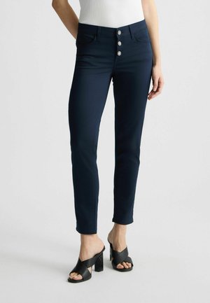 Pantaloni aderenti blu navy con una chiusura a cinque bottoni, due tasche laterali e una texture liscia. Abbinati a sandali neri con tacco a blocco.
