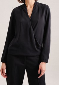 Blusa nera avvolgente realizzata in tessuto morbido, caratterizzata da un profondo scollo a V e maniche lunghe; include un dettaglio con un solo bottone. Abbinata a pantaloni neri.