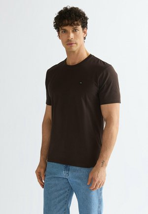 Uomo con capelli ricci che indossa una t-shirt marrone scuro semplice a maniche corte e jeans azzurri, in piedi davanti a uno sfondo bianco semplice.