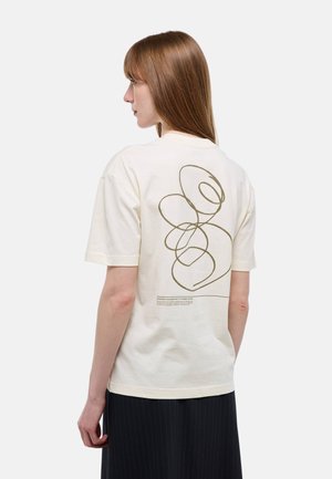 Femme aux cheveux châtain clair et raides, portant un t-shirt crème avec un dessin abstrait en lignes et un petit texte au dos, associé à un pantalon noir à fines rayures.