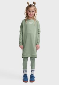 Jasnozielona sukienka-sweta z opadającymi ramionami i logo na piersi, w komplecie z jasnzielonymi legginsami w prążek i niebieskimi sneakersami.