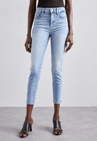 Jeans de tiro alto en azul claro con un ajuste slim y un largo recortado, que presentan un sutil desvanecimiento y un diseño clásico de cinco bolsillos. Tacones negros destalonados.
