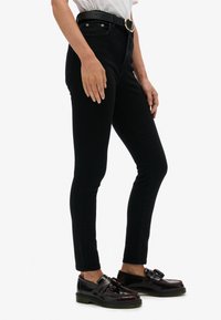 Vaqueros negros de mezclilla de talle alto con corte ajustado, que cuentan con bolsillos frontales y una hebilla de cinturón dorada, combinados con loafer negros brillantes.