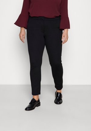 Kobieta ubrana w czarne dopasowane spodnie do kostek, czarne błyszczące eleganckie buty oraz bordową bluzkę z długim rękawem z rozszerzanymi mankietami.