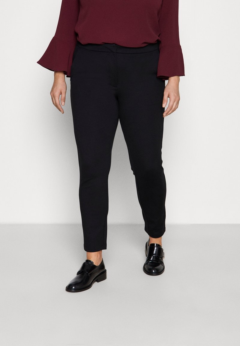 Femme portant un pantalon noir ajusté à la cheville, des chaussures de ville noires brillantes et un haut bordeaux à manches longues avec des poignets évasés.