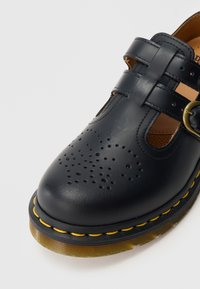 Sandale en cuir noir à bout fermé avec motif perforé, double brides, boucle en laiton et semelle en caoutchouc translucide avec couture jaune.
