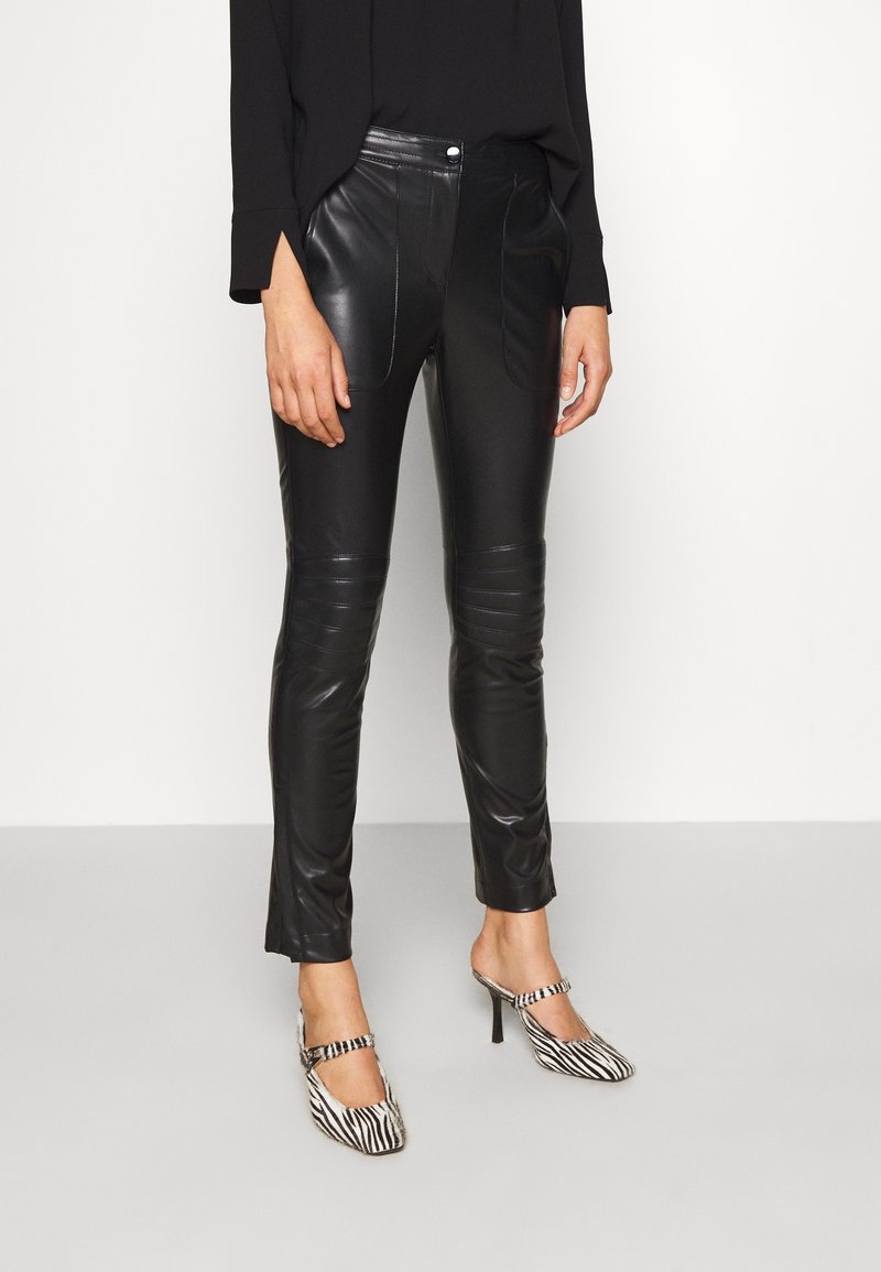 Pantaloni in faux pelle nera a vestibilità slim, con cuciture laterali e una texture sottile; indossati con scarpe con tacco a righe bianche e nere.