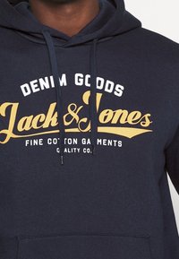 Námořnická modrá mikina z bavlny s bílým a zlatým textem: "DENIM GOODS Jack & Jones" a "FINE COTTON GARMENTS" a přední kapsou.