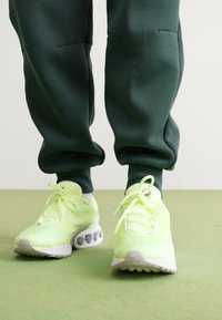 Neon sárga sportcipők hálós felsőrésszel, párnázott talppal és kerek sarokkal, sötétzöld jogger nadrággal egy világoszöld felületen.