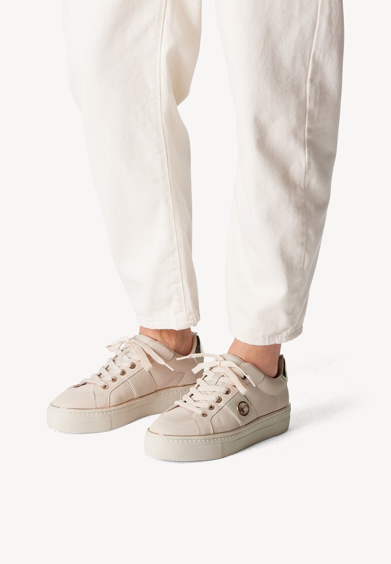 Beige sneakers featuring a smooth leather upper, lace-up design, subtle logo detail, and textured rubber sole, paired with white pants.