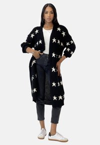 Cardigan noir en tricot avec des motifs d'étoiles blanches, ouverture à l'avant et manches longues, associé à un haut blanc et un jean noir, complété par des baskets blanches.