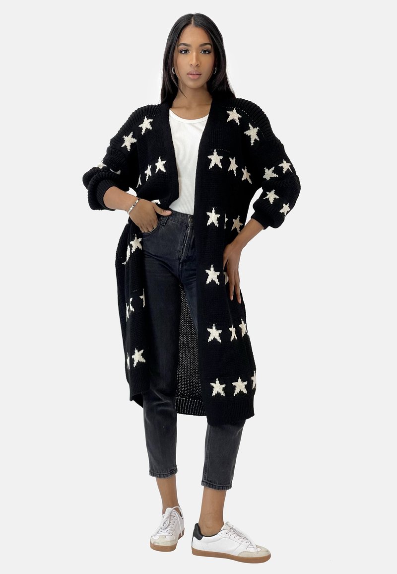 Cardigan noir en tricot avec des motifs d'étoiles blanches, ouverture à l'avant et manches longues, associé à un haut blanc et un jean noir, complété par des baskets blanches.