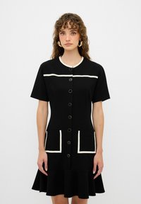 Vestido negro con mangas cortas, escote redondo, ribetes blancos en contraste, botones en el frente y dos bolsillos con parches bordeados de blanco. Falda acampanada.