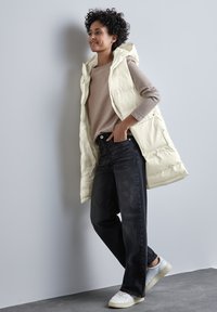 Světlobéžová péřová vesta s kapucí přes béžový svetr, v kombinaci s tmavými džíny a bílými teniskami, na šedém pozadí.