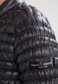 Zwarte pufferjack met een doorgestikt patroon, glanzende stof en geribbeld textuur. Heeft een logolabel op de mouw.