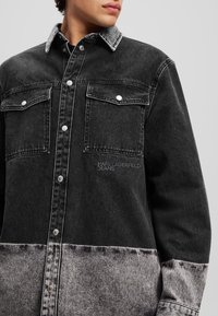 Sort denimjakke med kontrasterende grå bund, to brystlommer, hvid syning og metaltrykknapper. Har "KARL LAGERFELD JEANS" broderi.