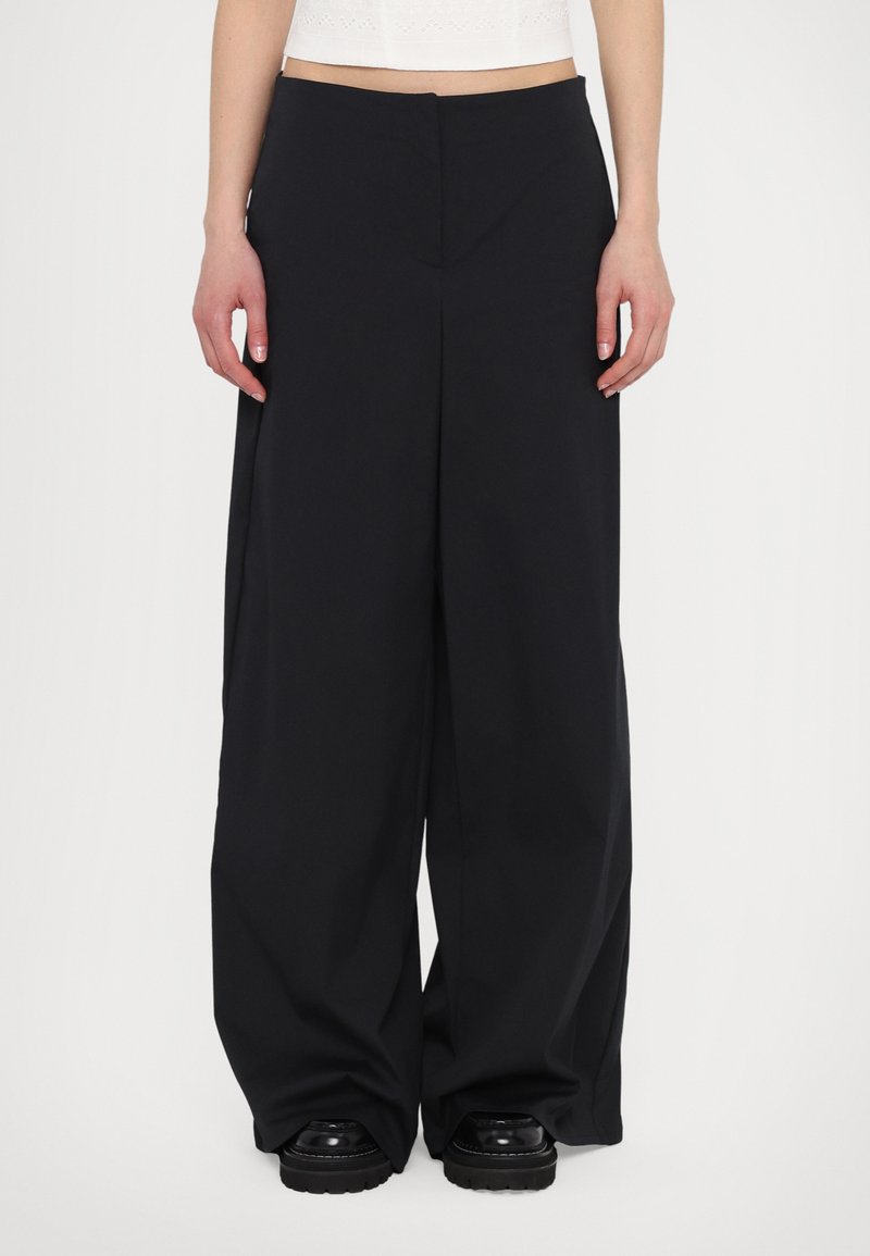 Wide-leg black trousers made of smooth fabric, featuring a tailored design with a pronounced front crease and side pockets.