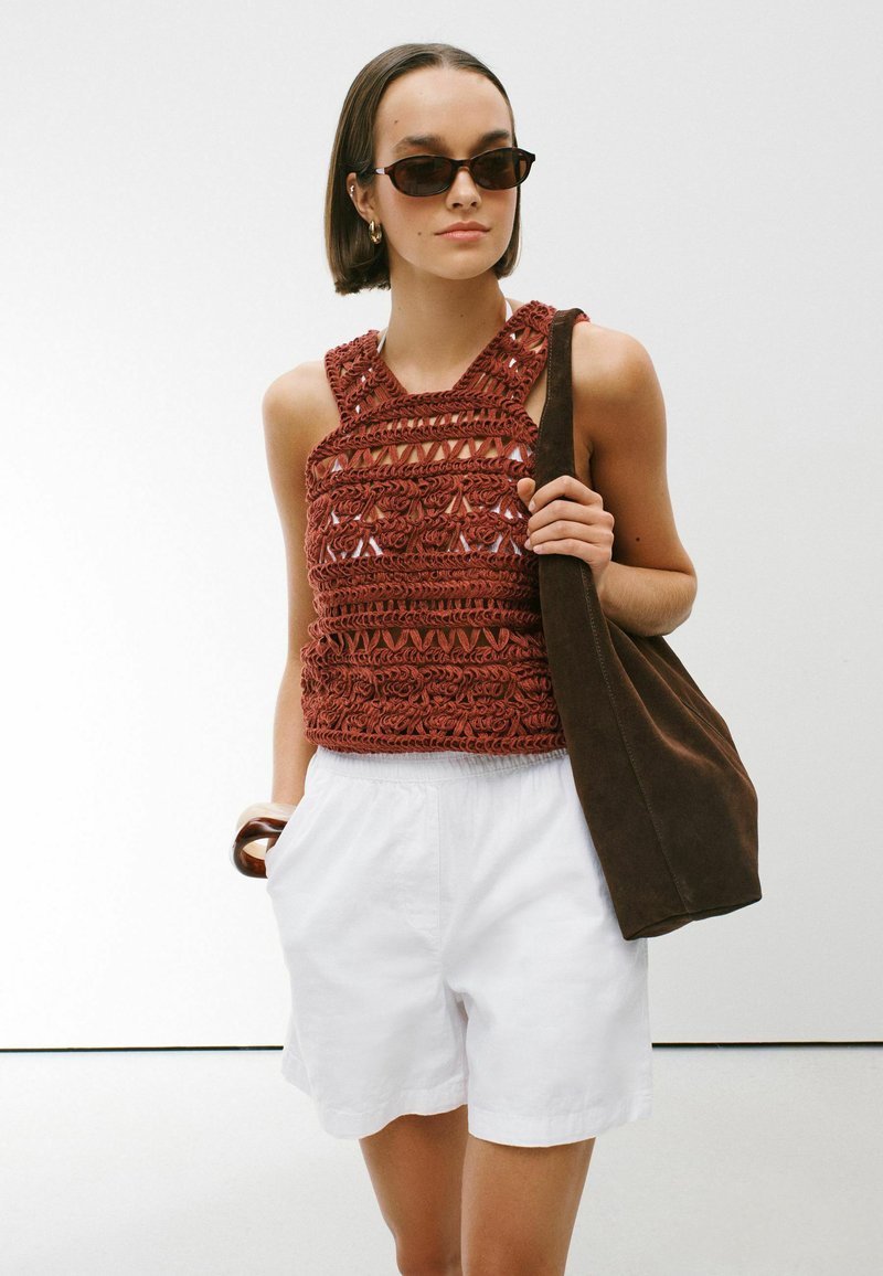 Woman wearing brown crocheted sleeveless top, white shorts, dark sunglasses, carrying a large brown shoulder bag and a wooden bracelet.