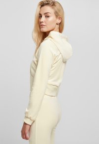 Hoodie coupé en tissu doux de couleur jaune pâle avec capuche à cordon, présentant des poignets ajustés et une texture lisse.