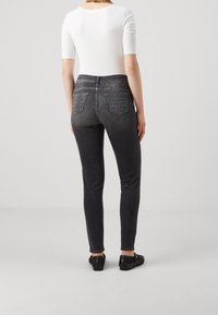 Jeans ajustados de mezclilla gris oscuro con dos bolsillos traseros que tienen patrones de costura geométricos, combinados con una blusa blanca ceñida y zapatos negros.