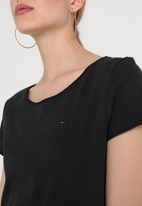 Černý bavlněný top s krátkým rukávem, kulatým výstřihem a malým logem. Modelka má na sobě velké kruhové náušnice. Hladká textura.