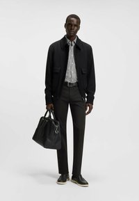 Black bomber jacket with two front pockets, striped shirt underneath, dark trousers, and black leather shoes with a contrasting sole. Holding a black bag.