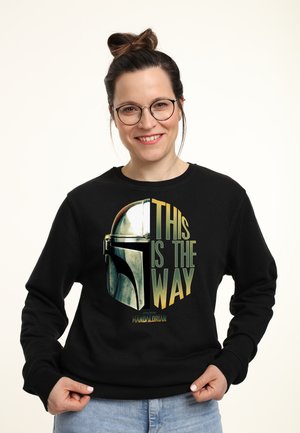 Kvinde iført briller og en sort sweatshirt med en Mandalorian hjelm og teksten "This is the way" i gult og grønt.