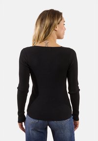 Donna con lunghi capelli biondi che indossa un top a maniche lunghe nero a coste e jeans blu, vista di spalle, che mostra un tatuaggio sul collo.