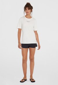 Weißes Baumwoll-T-Shirt mit kurzen Ärmeln und rundem Ausschnitt, mit einem schwarzen Textlogo. Kombiniert mit schwarzen Shorts und schwarzen Flip-Flops.
