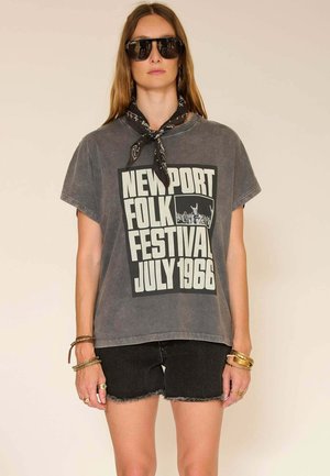 Femme portant un t-shirt gris du Newport Folk Festival de juillet 1966, un short en denim noir, un foulard noir noué autour du cou, de grandes lunettes de soleil noires et des bracelets dorés.