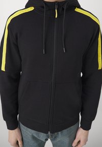 Černá mikina na zip s žlutými pruhy značky Emporio Armani EA7 na ramenou, kterou nosí osoba v modrých džínách, ruce viditelné po stranách.