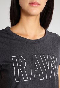 Uhlově šedé tričko vyrobené z bavlny, s velkým bílým potiskem "RAW" na přední straně. Krátké rukávy, kulatý výstřih, měkká textura.