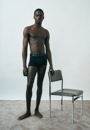 Mężczyzna stoi boso na dywanie, nosi czarną bieliznę Tommy Hilfiger, opierając rękę na szarym metalowym krześle z tapicerowanym siedziskiem i oparciem.