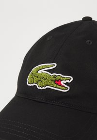 Fekete baseballsapka textúrált zöld krokodil logóval és piros kiegészítőkkel, fehér körvonallal a logón és ívelt árnyékkal.