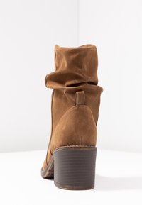 Bottine en daim marron avec une tige ample et un bout arrondi. Elle présente un talon moyen empilé et une petite languette à l'arrière pour un enfilage facile.