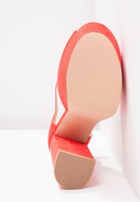 Chaussure à talon haut en suède rouge avec un talon épais, une semelle extérieure lisse de couleur nude et un design latéral ouvert. Le talon présente une surface texturée.
